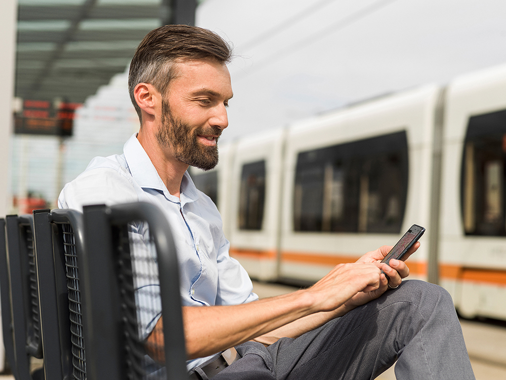 Mann sitzt auf Bank mit Smartphone in der Hand und Straßenbahn im Hintergrund