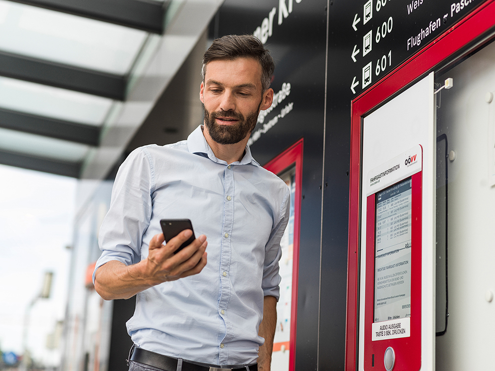 Mann steht an einer Straßenbahnhaltestelle und wirft einen Blick auf sein Smartphone.